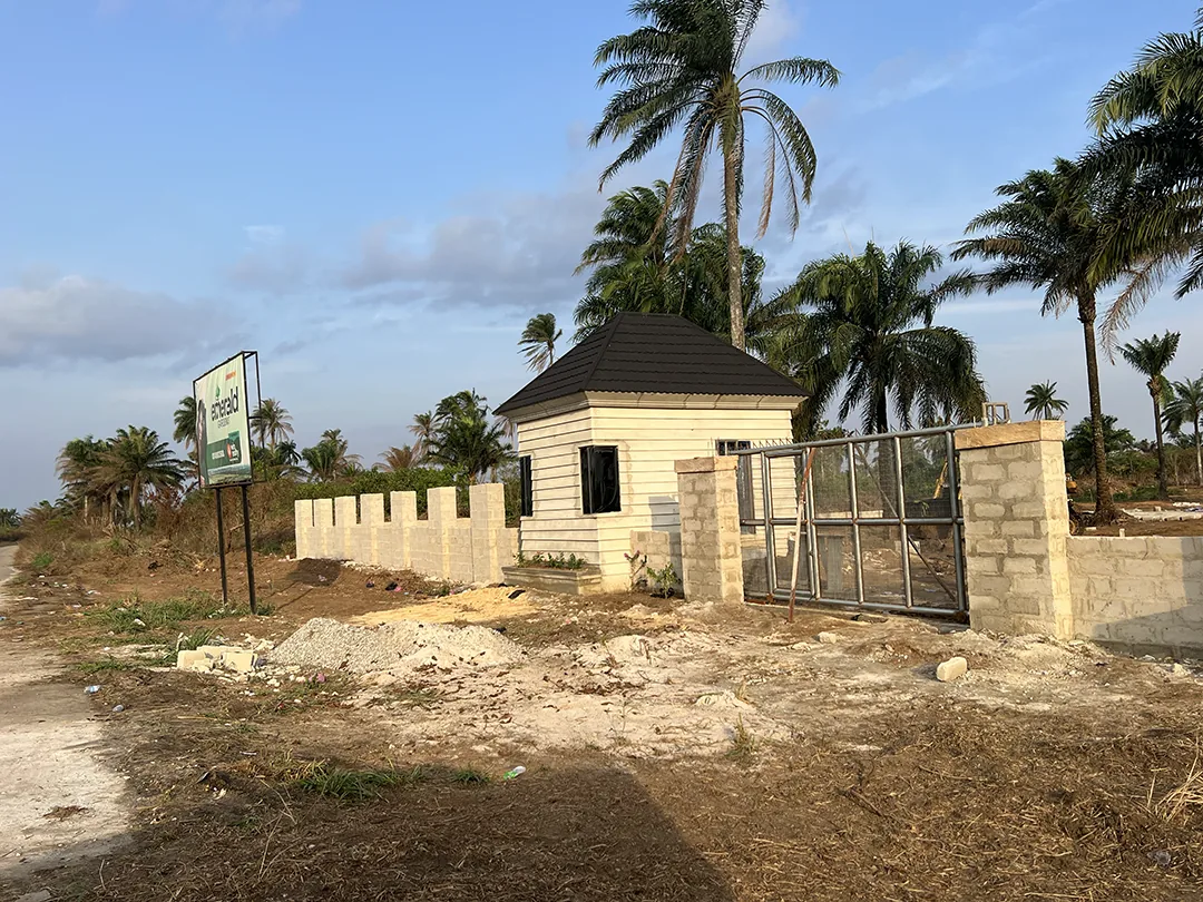 Emerald Gardens in Ozuzu-Etche: entrance showing its gatehouse, metal gate, boundary wall and palm trees.